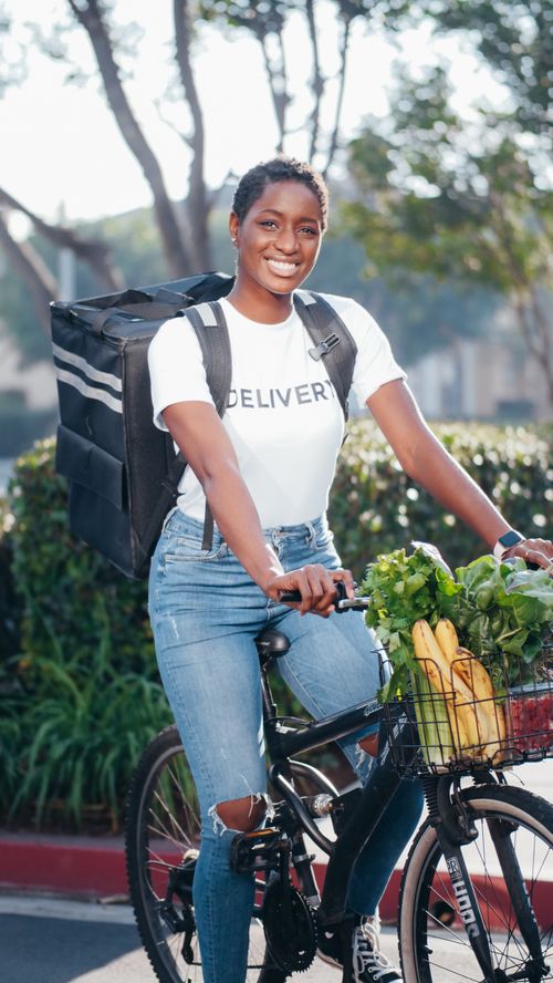Free A cheerful delivery rider on a bicycle carrying fresh groceries in an outdoor setting. Stock Photo