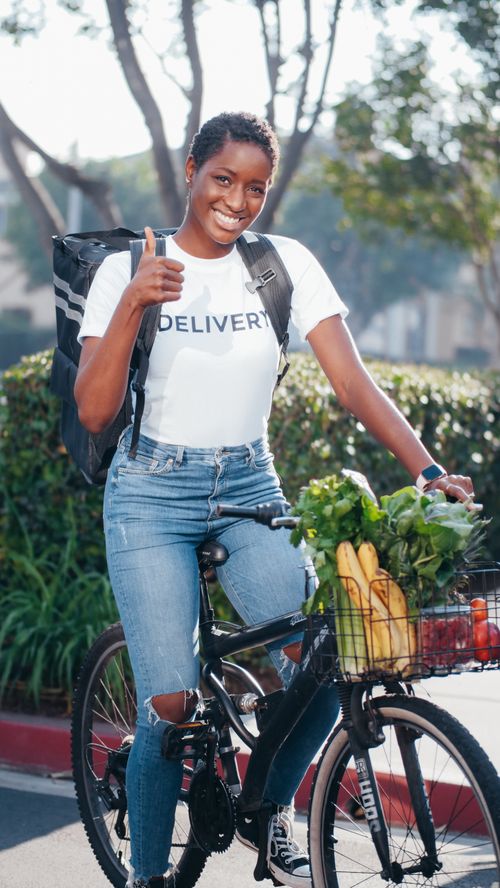 Free Smiling African American woman on bicycle delivering groceries outdoors, showing thumbs up. Stock Photo