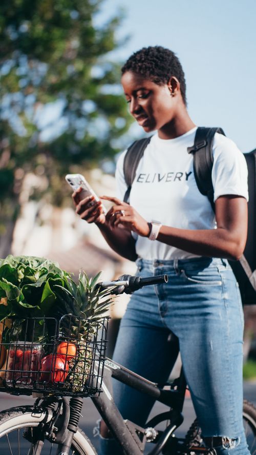 Free African American woman checking phone while delivering groceries on a bicycle outdoors. Stock Photo