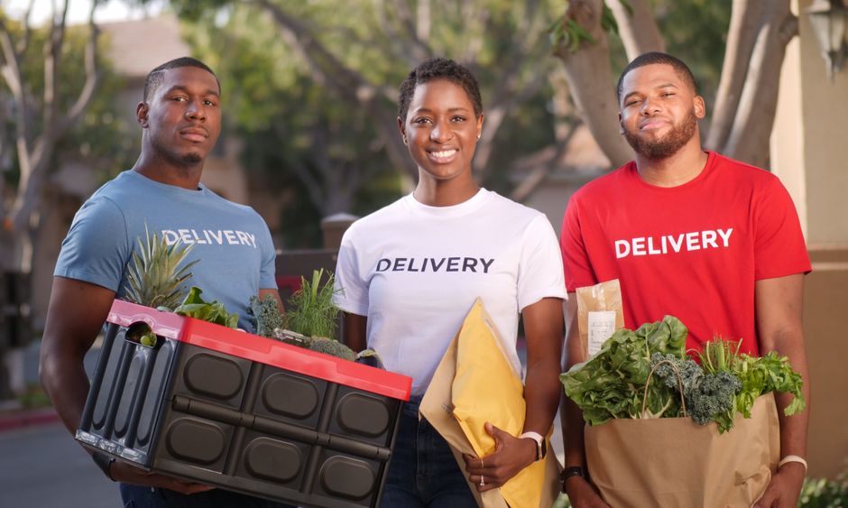 Dark-skinned Nigerian delivery worker in a branded vest handing over a box of groceries at the door of a modest Nigerian home to a smiling elderly Black Nigerian woman in a floral housedress, bright afternoon light, photorealistic