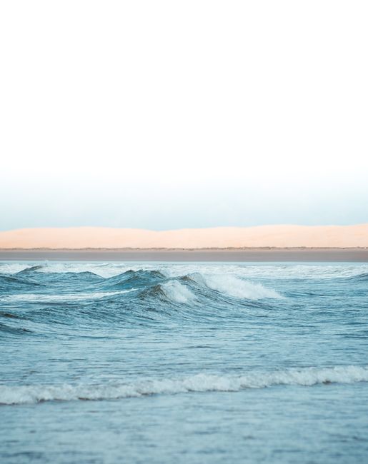 Tranquil ocean scene with blue waves and distant sandy dunes under a clear sky.