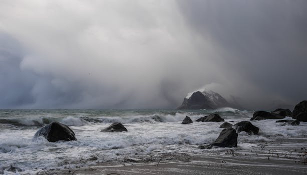 Waves crash against dark rocks under a moody, cloud-filled sky at a rugged seashore.