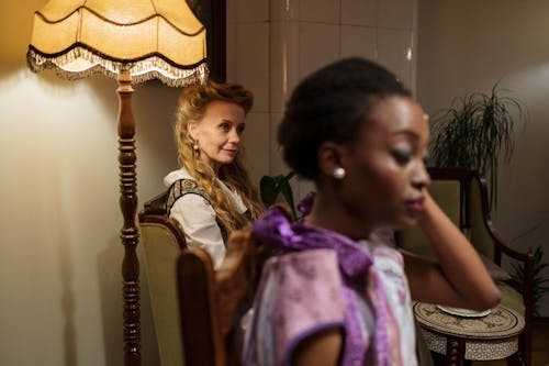 Two women dressed elegantly, sitting in a classic indoor setting with warm lighting.