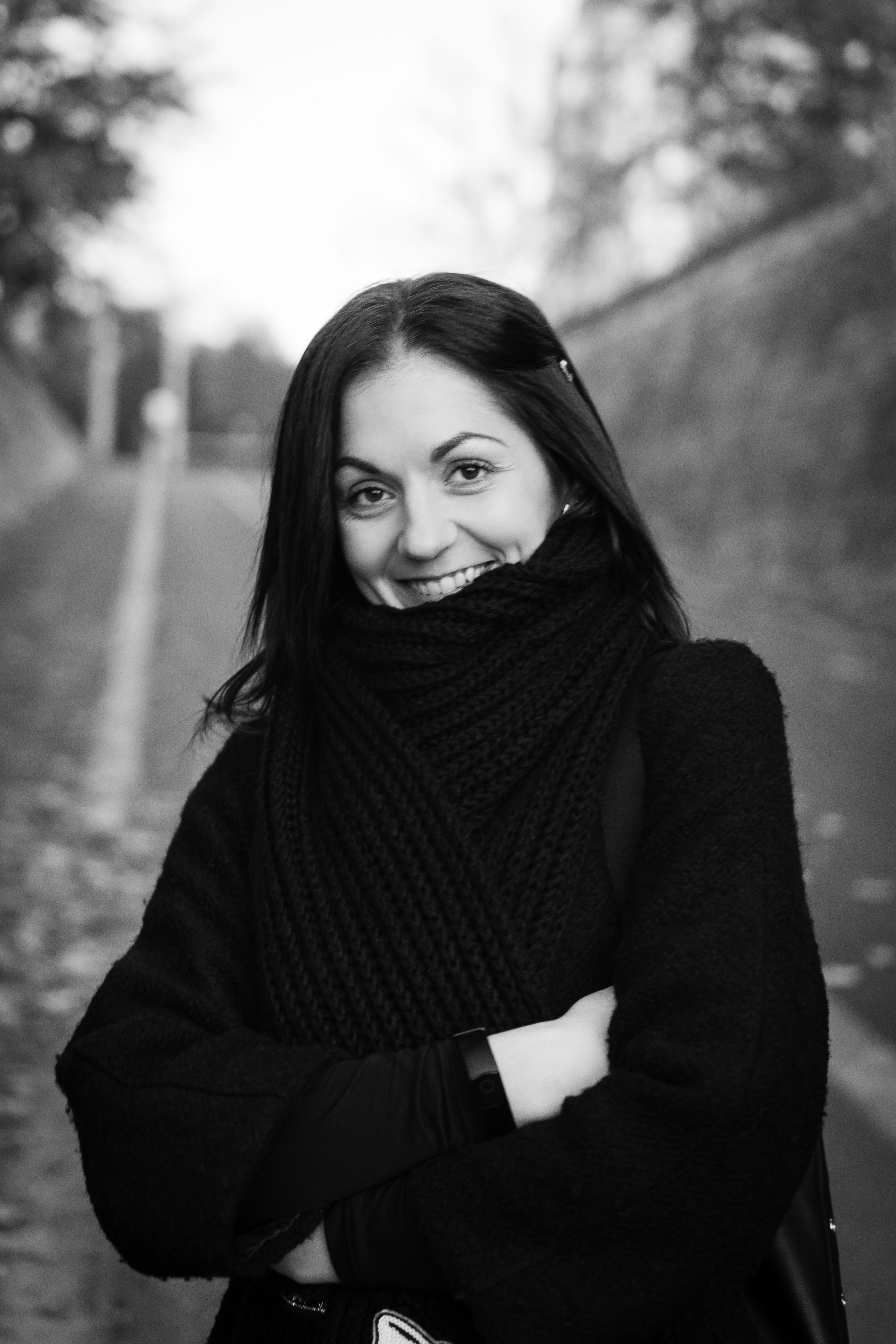Portrait of a woman in a black scarf and jacket smiling outdoors on a fall day.