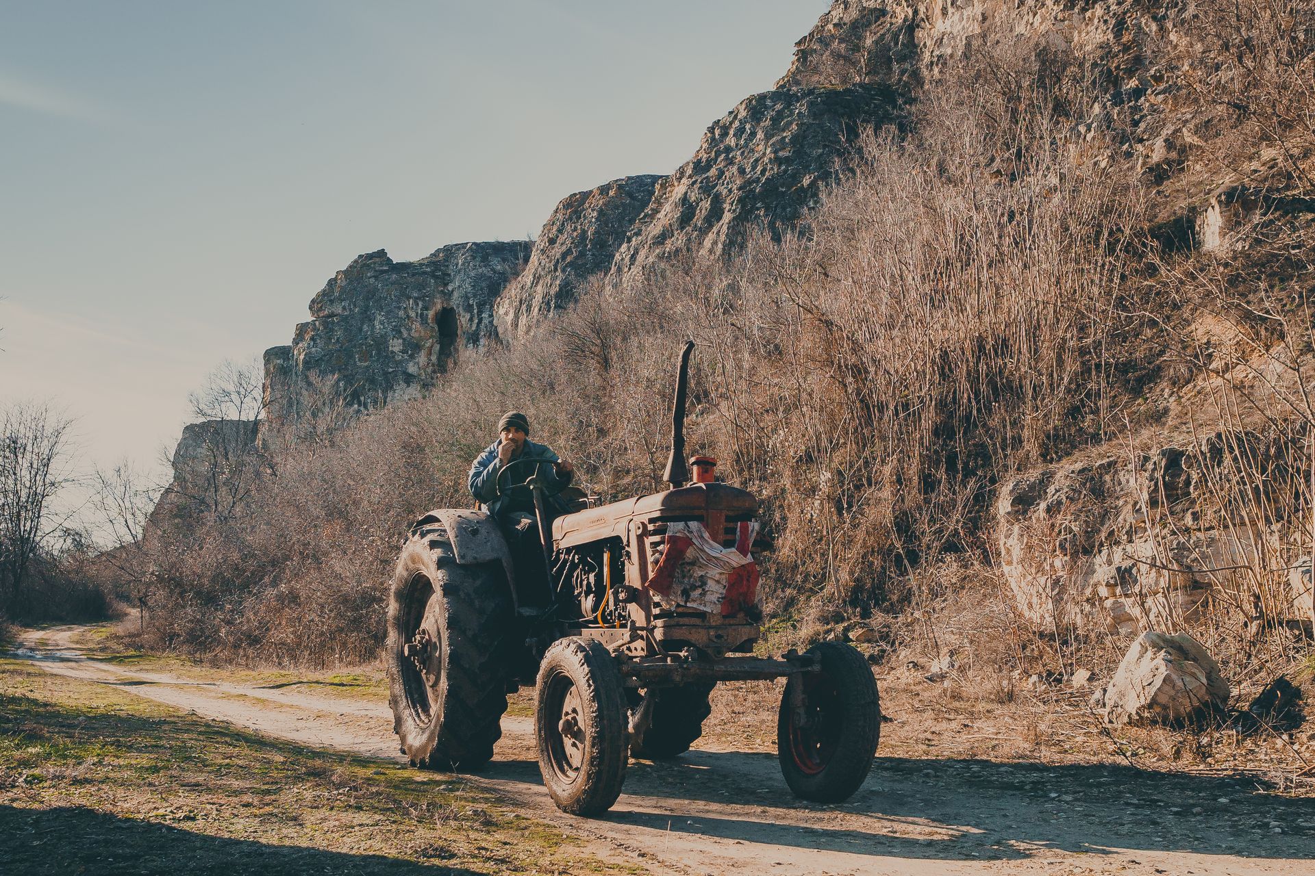 A man drives a vintage tractor along a mountain dirt road in a rural setting under clear skies.