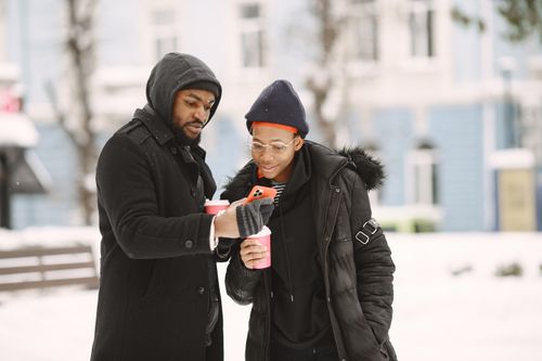 Free An African American couple dressed in winter jackets enjoying hot drinks outdoors while looking at a smartphone on a snowy day. Stock Photo