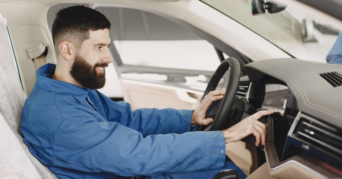 Mechanic in blue coveralls interacts with car dashboard, smiling and focused.