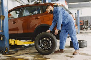 Man in Blue Coveralls Standing Beside an Orange Car Checking on the Tire