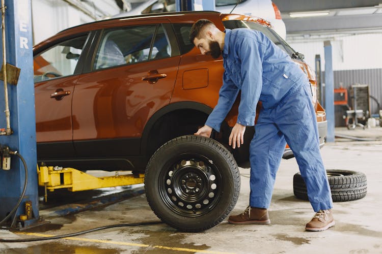 Man In Blue Coveralls Standing Beside An Orange Car Checking On The Tire