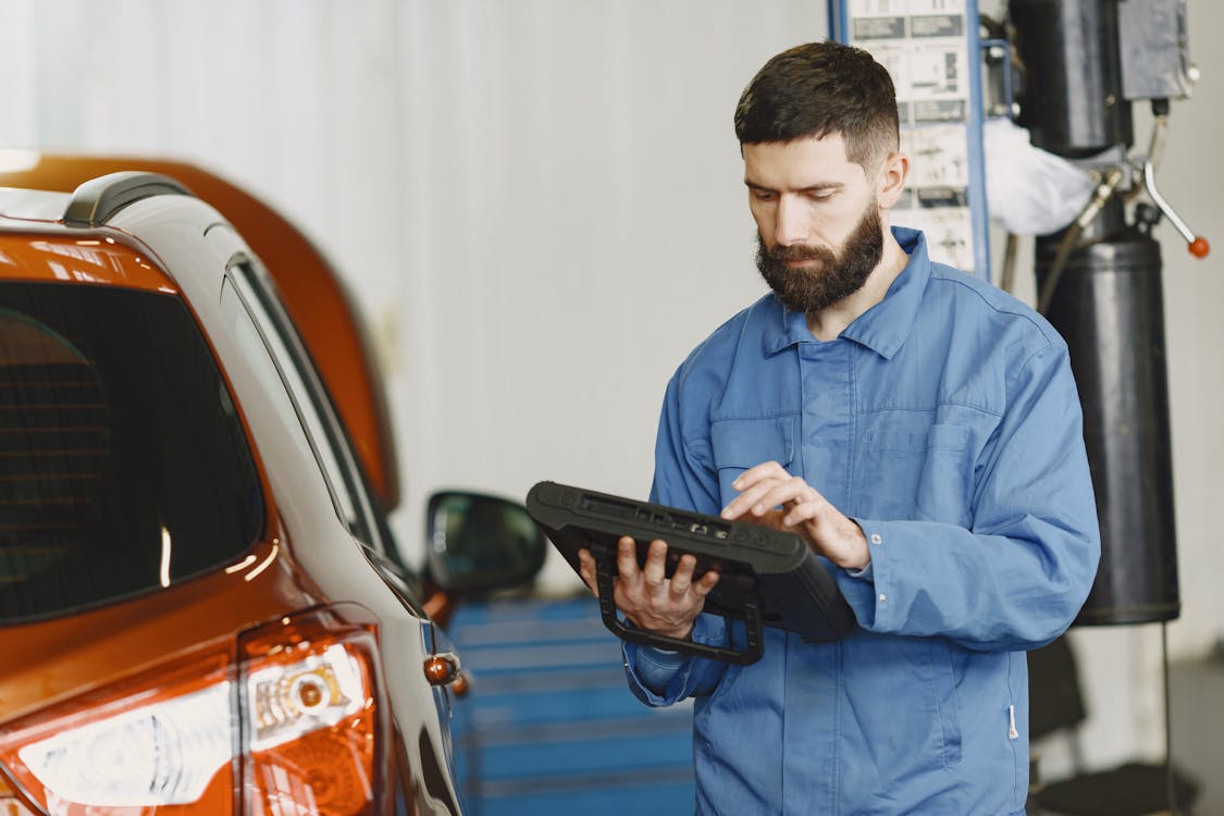 Free Man in Blue Coveralls Standing Beside an Orange Car Using an Automotive Diagnostic Tool Stock Photo