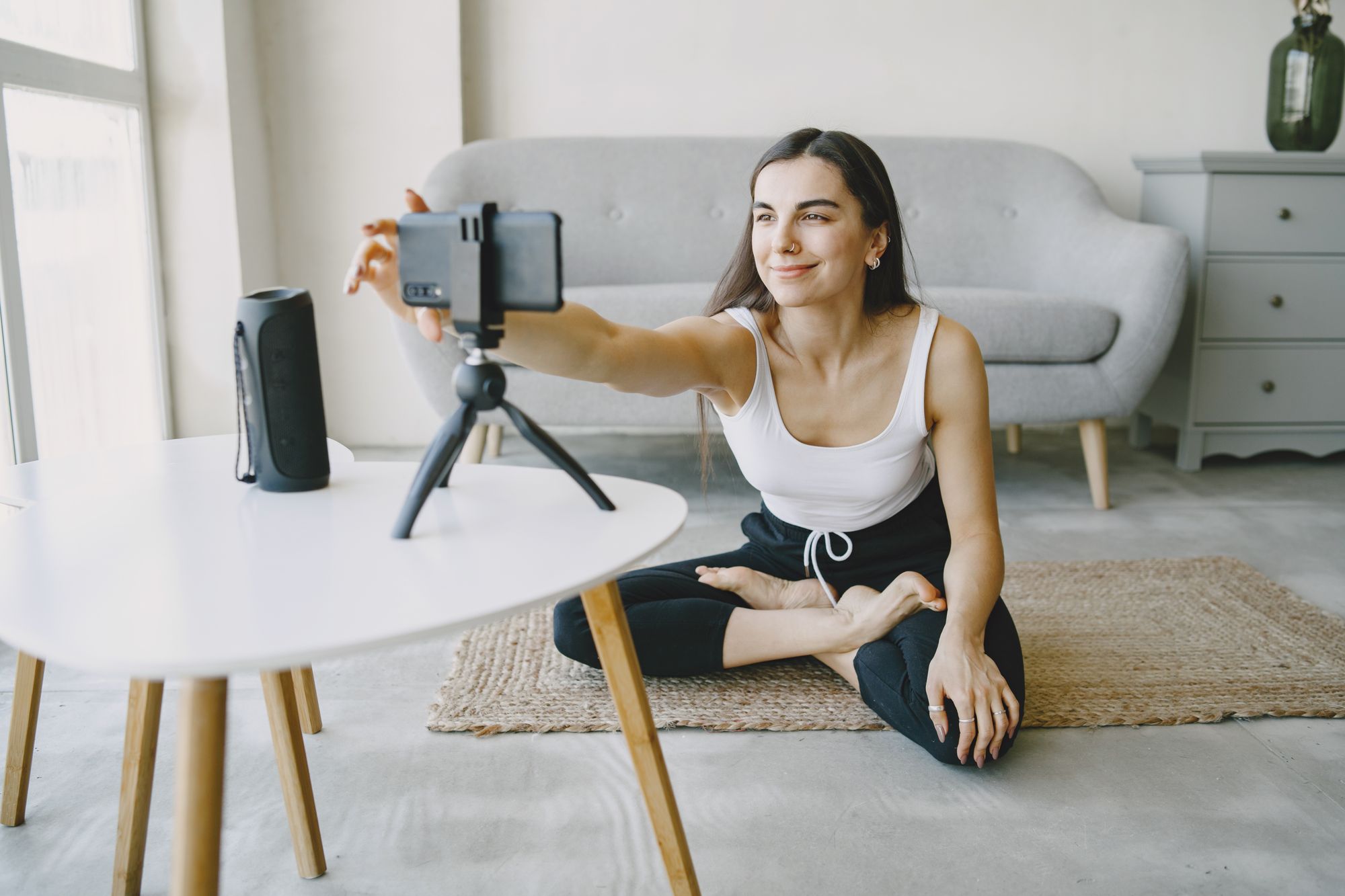 Smiling woman in a tank top vlogging indoors using a smartphone and tripod setup.