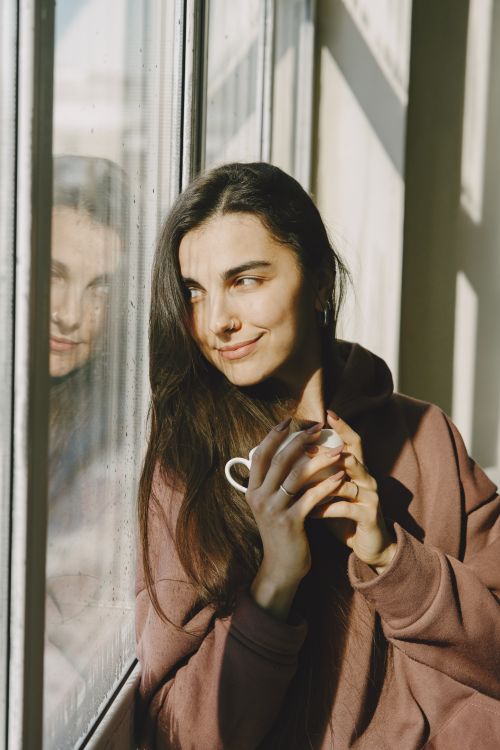 Free Young woman in cozy hoodie enjoying sunlight by a window, holding a cup, reflecting calmness and warmth. Stock Photo