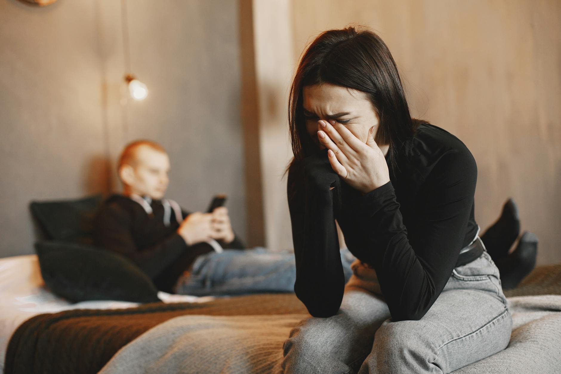 Woman crying on bed, man in background, illustrating relationship stress.