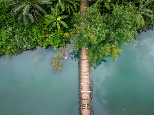 Free Aerial view of a rustic wooden footbridge crossing a tranquil turquoise river surrounded by lush green vegetation. Stock Photo