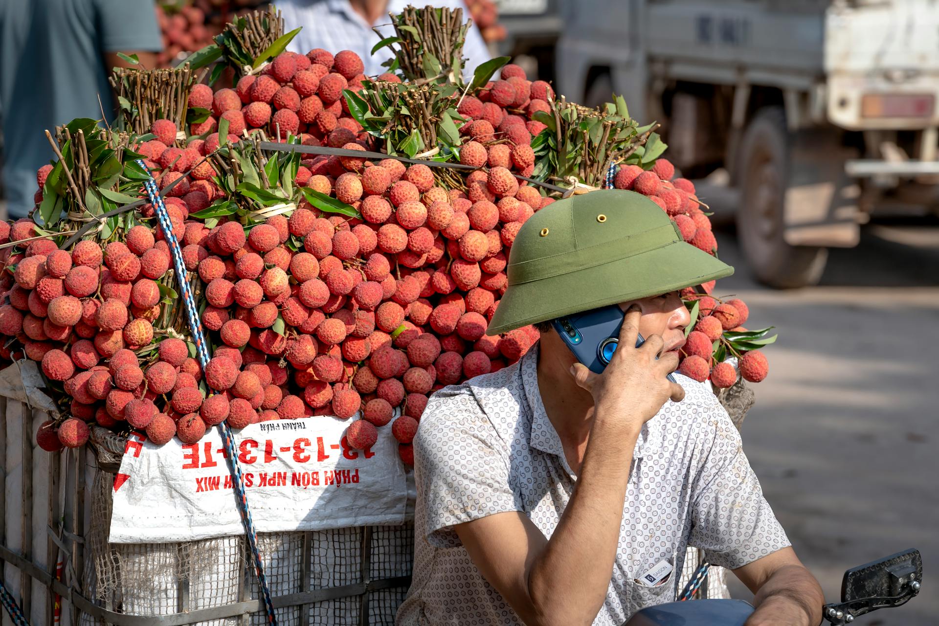 Asian vendor on phone selling fresh lychees at an outdoor market with a large fruit display.