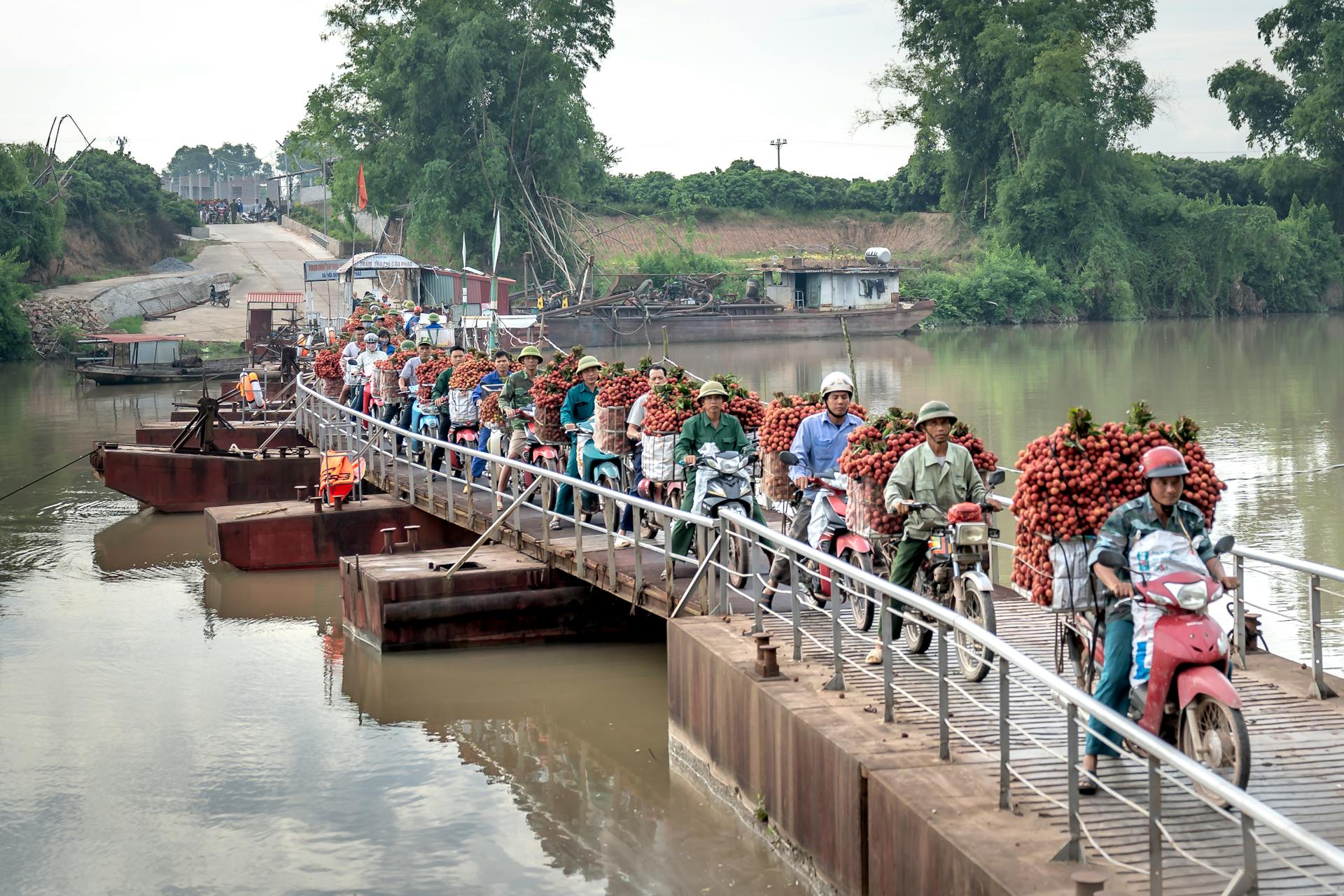 Motorbike vendors carrying fruit baskets across a rural footbridge over a river.