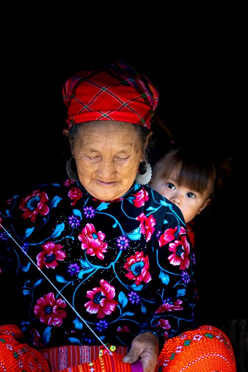 Free A senior woman sewing in colorful attire with a child peeking from behind. Stock Photo