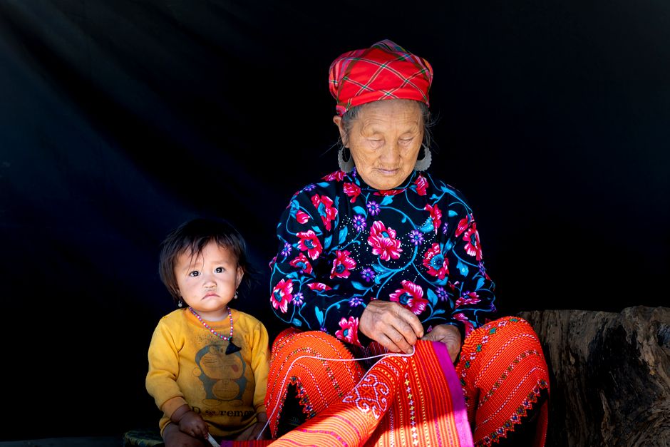 An elderly woman wearing traditional clothing sews with a young child sitting beside her, showcasing cultural heritage.