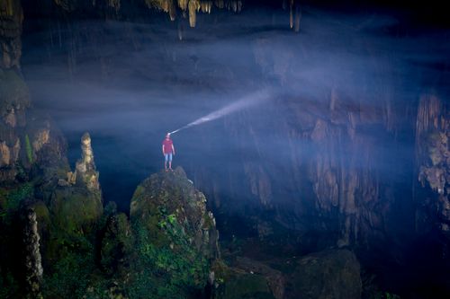 Free A person stands on a cave rock, illuminating the vast interior with a headlamp, showcasing adventure. Stock Photo