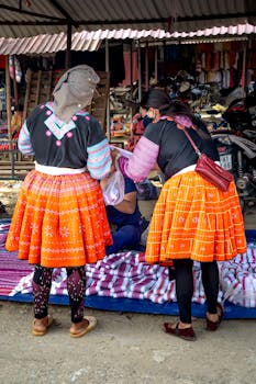 Two women in traditional wear shopping at a colorful market stall displaying textiles.