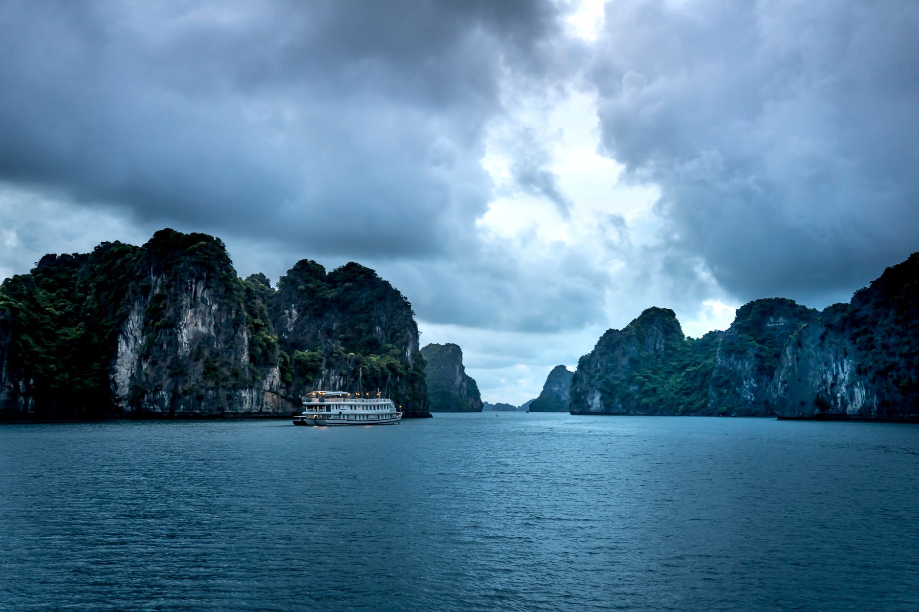 Dramatic view of a cruise ship sailing in Halong Bay's limestone cliffs under cloudy skies.