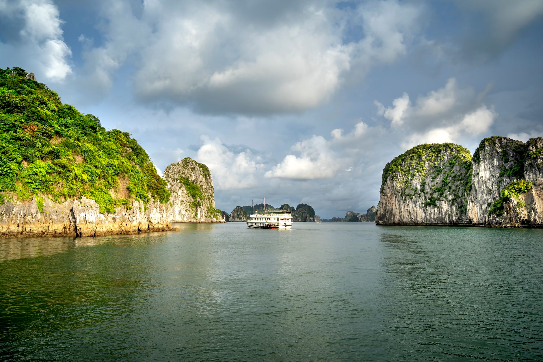 Erkunden Sie die atemberaubenden Felsformationen und ruhigen Gewässer der Ha Long Bay, mit einem Kreuzfahrtschiff unter einem dramatischen Himmel.