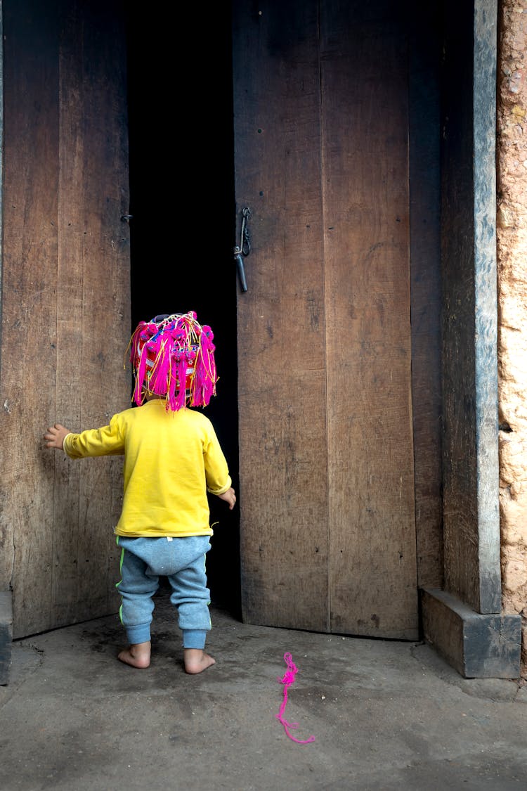 Little Child With A Bright Pink Hat Standing In Front Of Wooden Front Door 