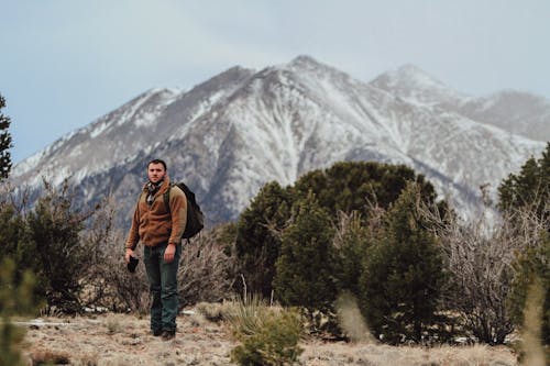 A Man in Brown Jacket Standing on Brown Field with a Snow Covered Mountain on the Background