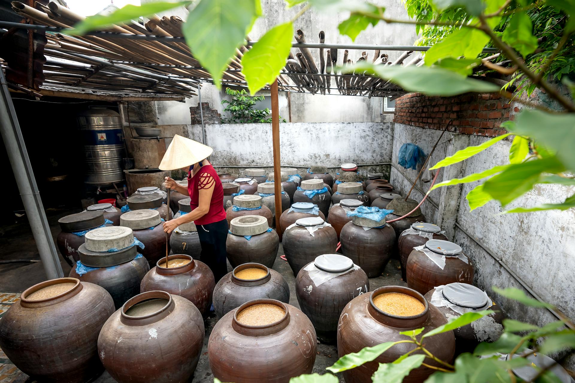 Person in Conical Hat Preparing Traditional Food in Big Jars