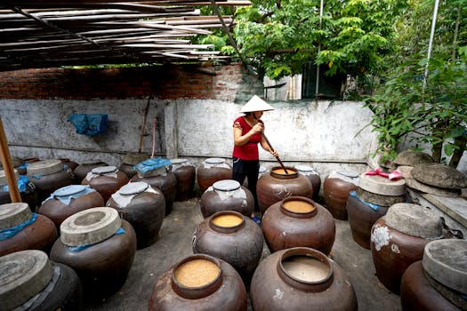 A person stirring contents in traditional jars outdoors, wearing a conical hat.