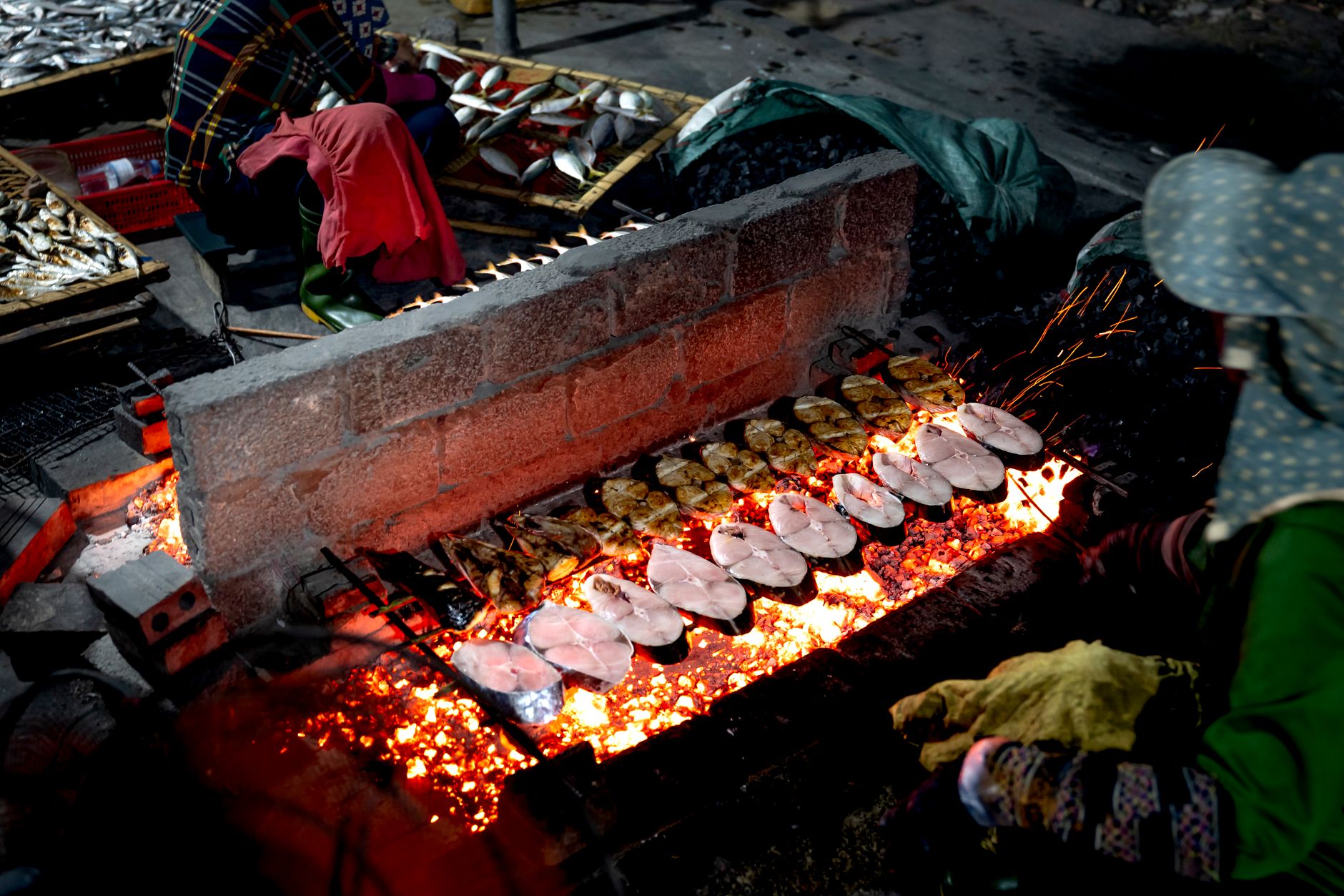 Fresh fish being grilled over open flames in a bustling street market by local vendors at night.