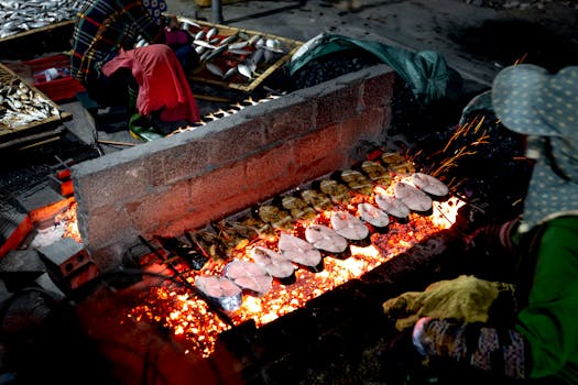 Fresh fish being grilled over open flames in a bustling street market by local vendors at night.