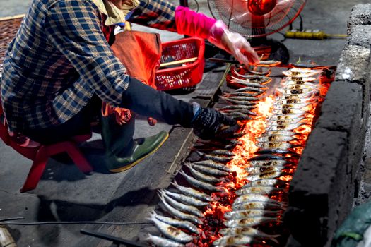 A vendor grills fish on an open fire, captured in an indoor market setting.