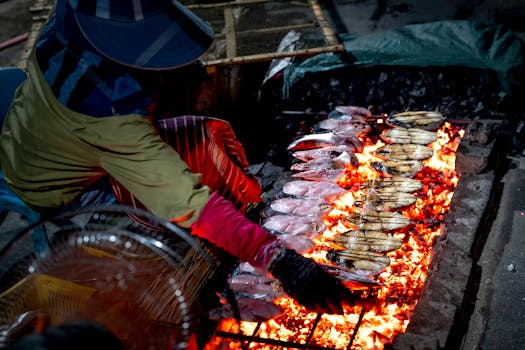 Street vendor grilling fish over open fire at night market.