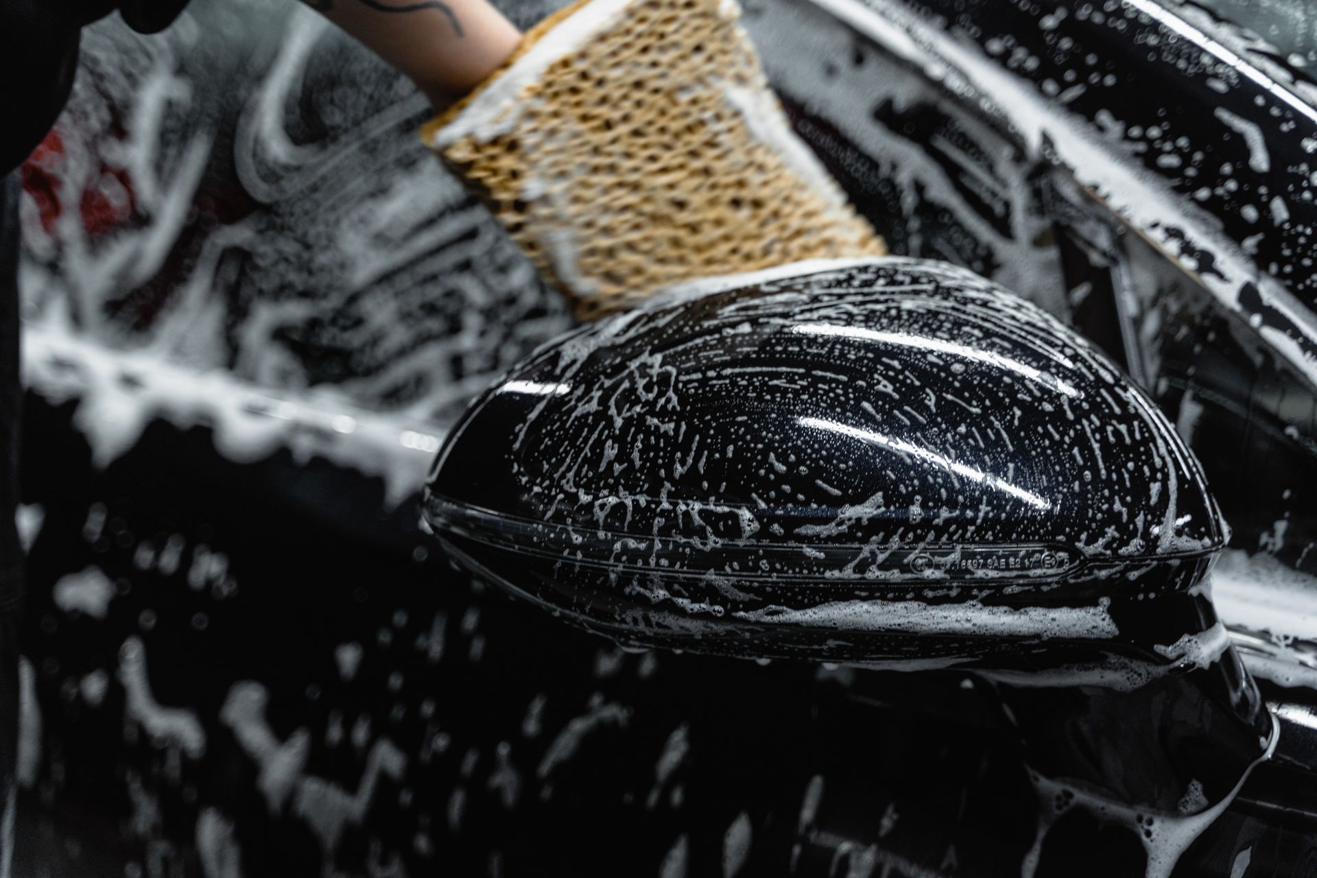 Hand scrubbing a car mirror with a sponge covered in soap at a car wash.