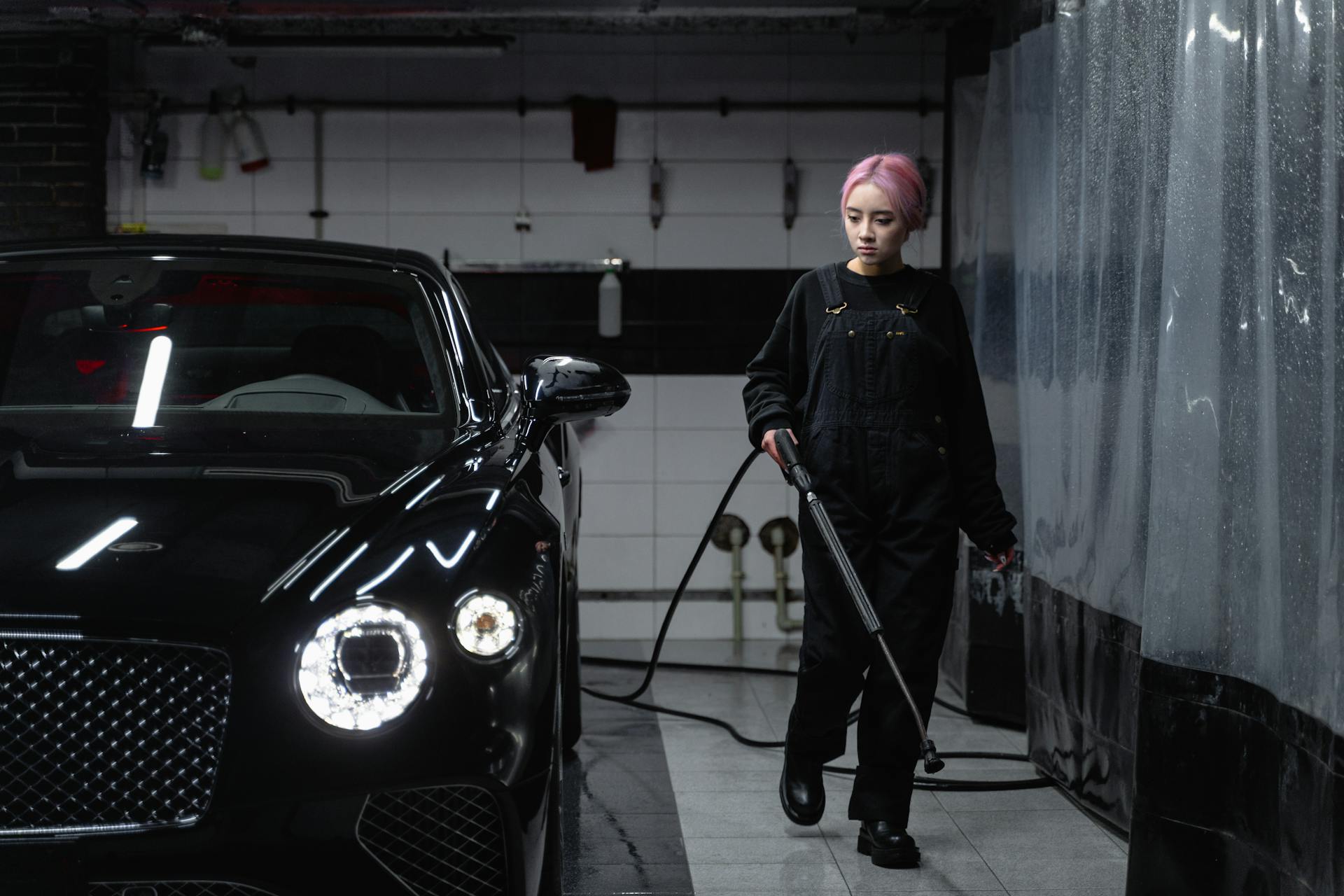 Woman cleaning luxury black car at car wash garage using hosepipe.