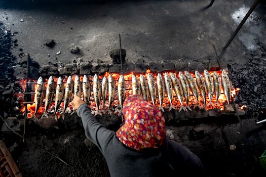 A person grills fish on a street in a traditional style over an open charcoal fire.
