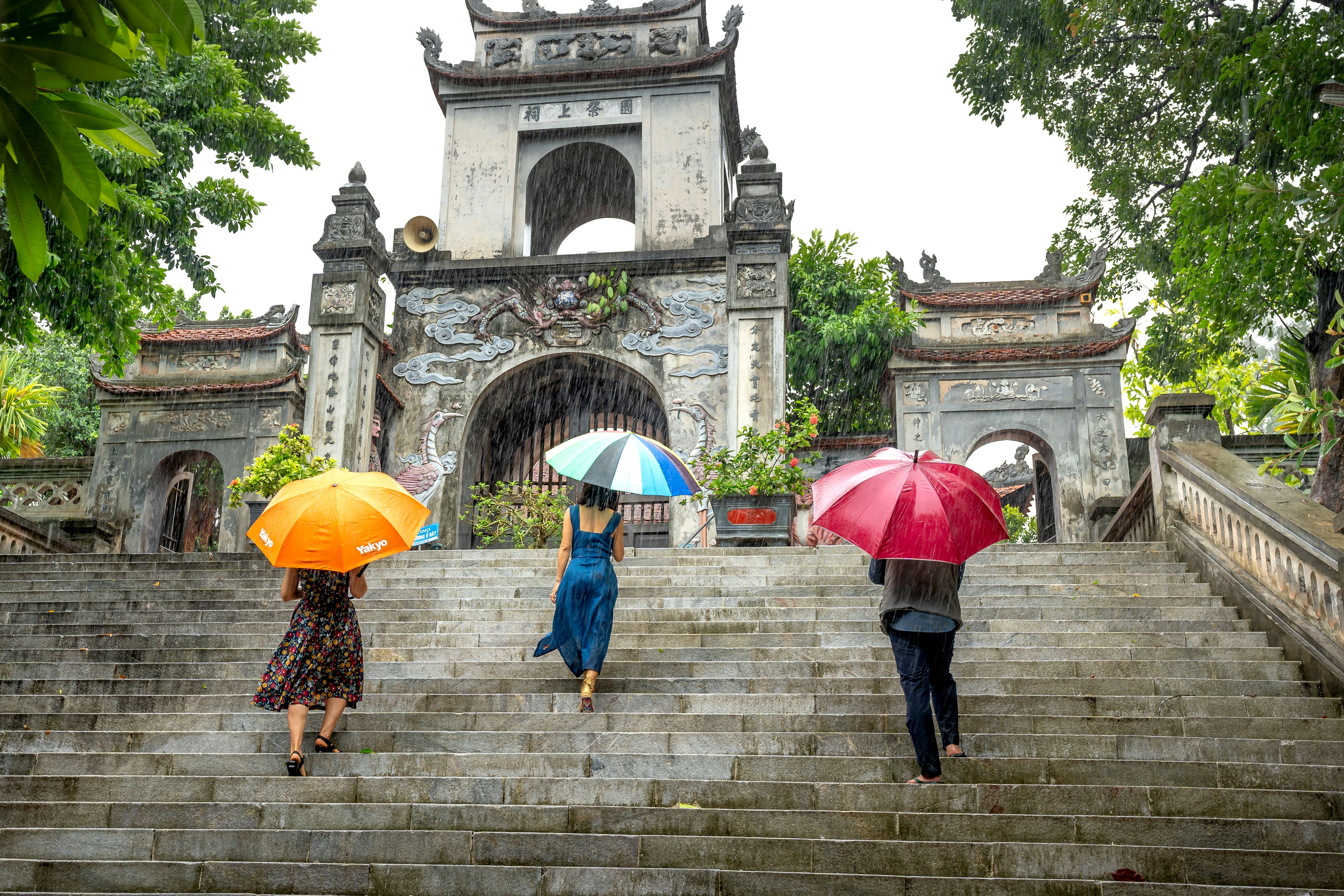 Three individuals with colorful umbrellas climb stone steps under rain towards a historic gate.