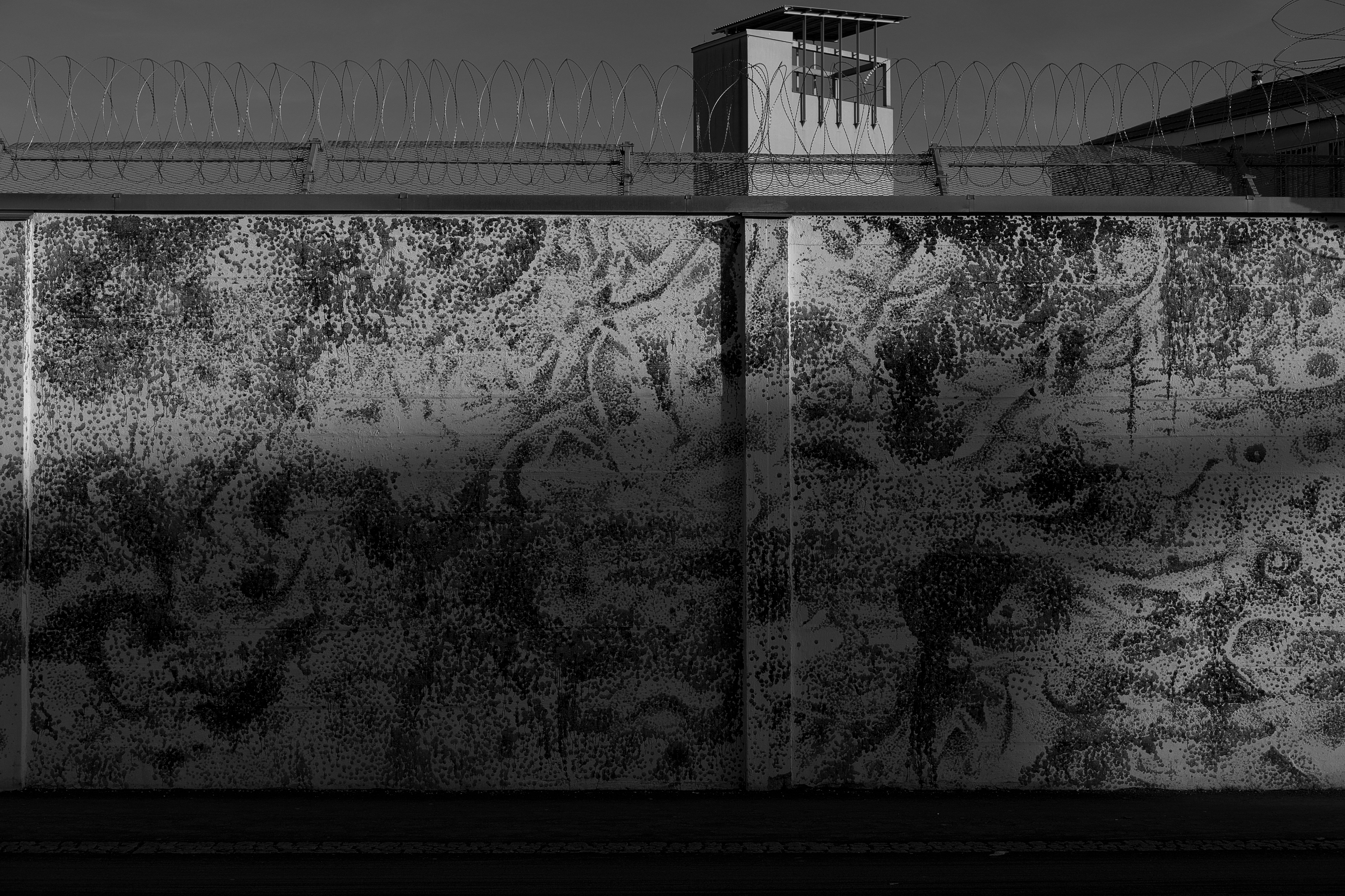 Black and white photo of a textured wall with barbed wire and a guard tower in shadow.
