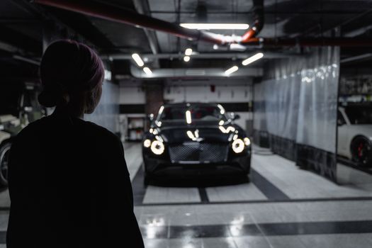 A silhouette of a person admiring a luxurious black car in a dimly lit indoor garage.