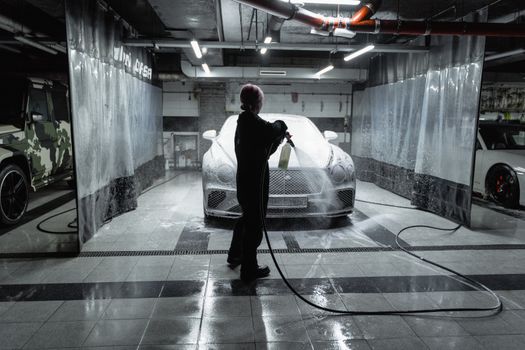A person using a hose to spray a car with snow foam in an indoor carwash setting.