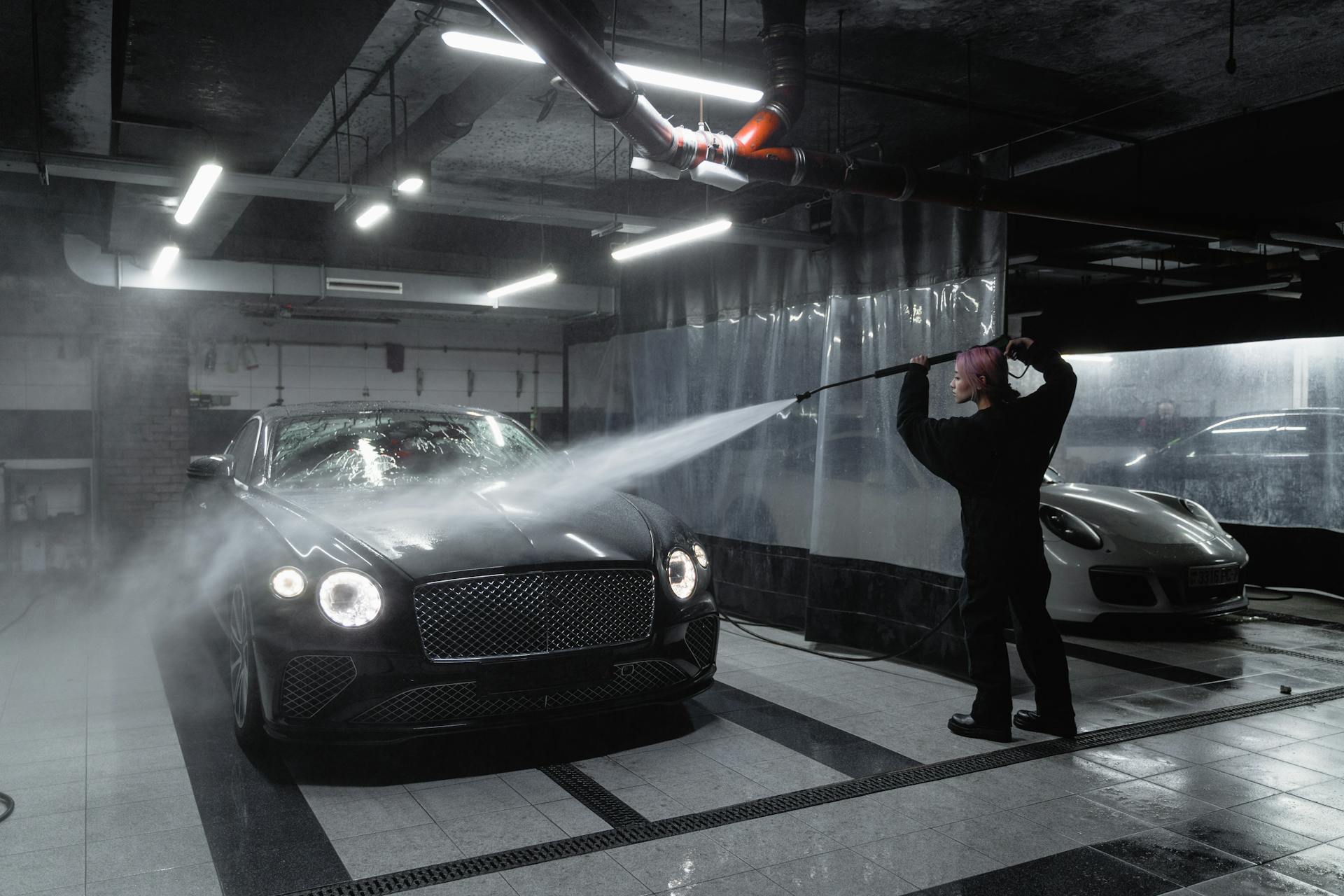 A person washing a black luxury car in a dimly lit garage using a pressure washer.