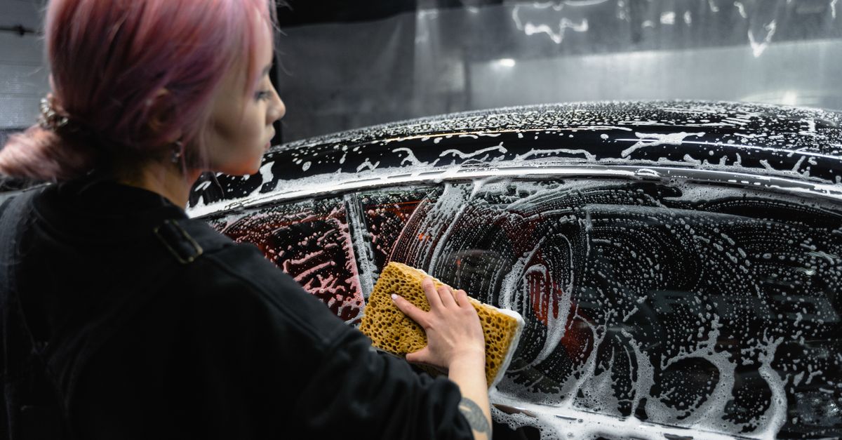 Woman washing black car with sponge, creating foamy soap suds indoors.