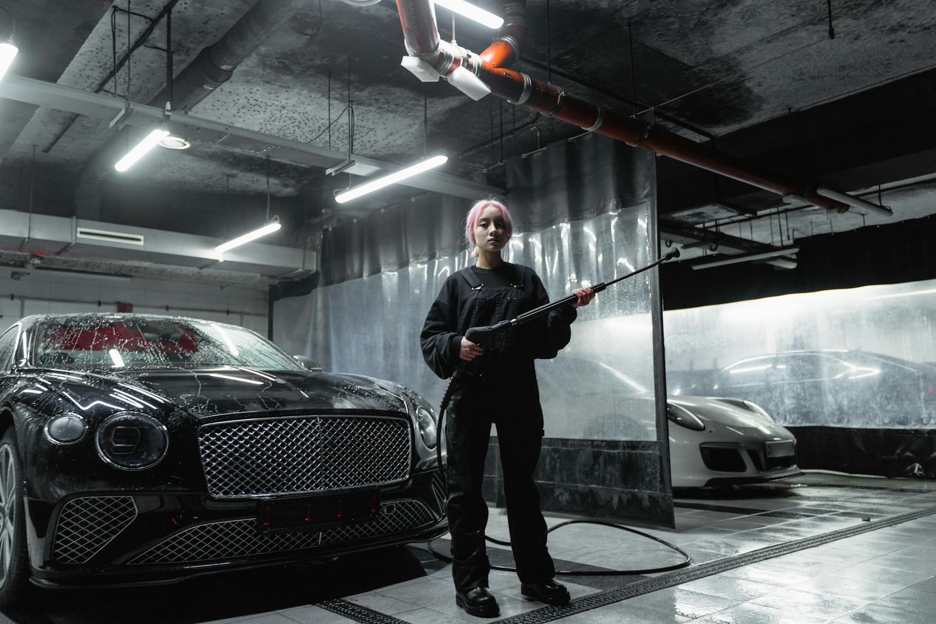 A woman in a car wash space using a power washer to clean luxury cars, captured indoors.