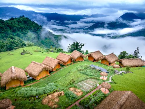 Free Scenic aerial view of straw-roofed huts nestled in lush green highlands with misty mountains. Stock Photo