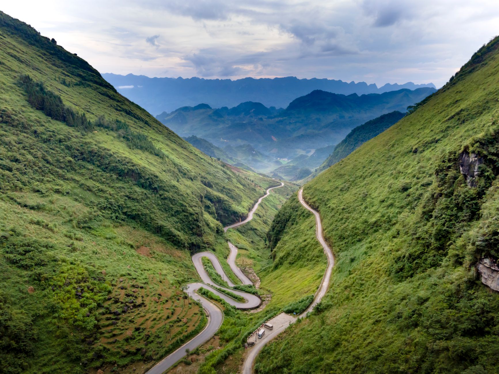 Aerial view of a winding road through a verdant mountainous landscape under a cloudy sky.