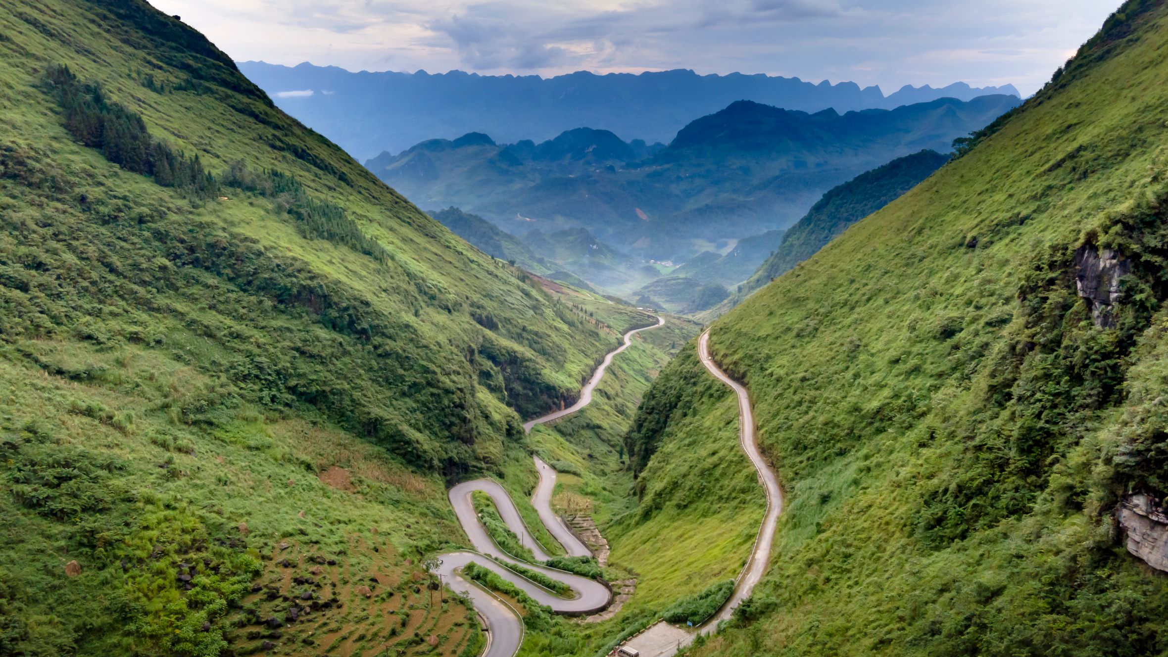 Aerial view of a winding road through a verdant mountainous landscape under a cloudy sky.