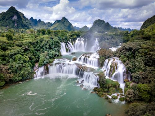 Free A picturesque multi-tiered waterfall cascading through a vibrant mountain landscape under a cloudy sky. Stock Photo
