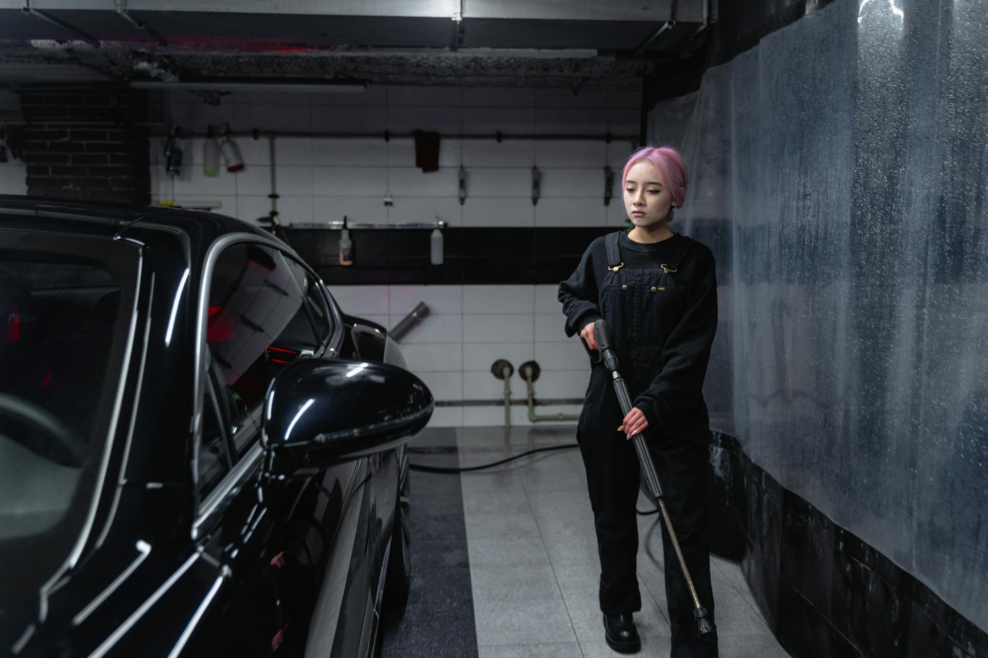 An adult woman with dyed hair using a pressure washer in an indoor car wash.