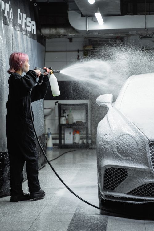 Free Young woman using a pressure hose to spray foam on a car in a garage setting. Stock Photo